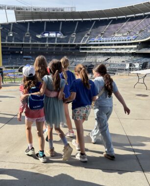 A group of six girls walk arm in arm across a walkway in what appears to be a baseball stadium.