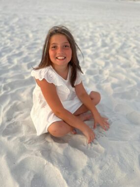 A young girl sits on the sand at the beach. She's wearing a white dress and smiling happily at the camera.